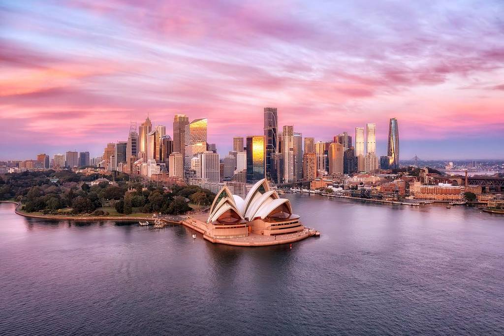 Sydney Opera House by the water at sunset, with city skyscrapers behind it and a pink-purple sky