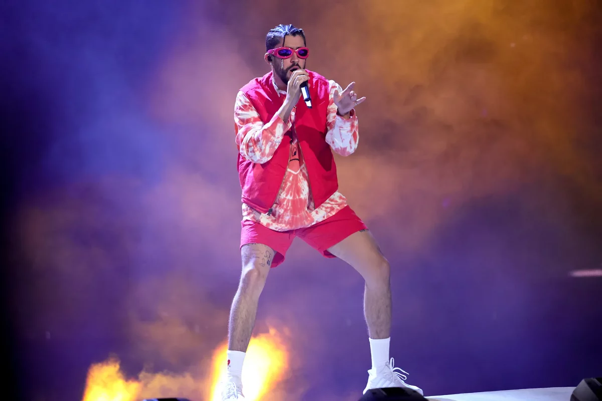 Bad Bunny performs on stage in a red vest and pink sunglasses, singing into a microphone amid smoke and colourful concert lighting