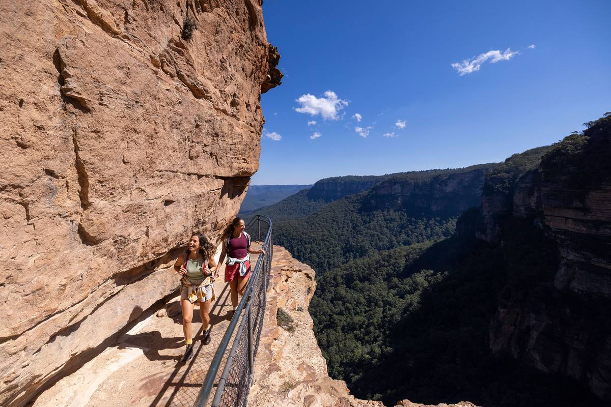 Hikers walking along a narrow clifftop trail with sandstone walls and sweeping Jamison Valley views in the Blue Mountains