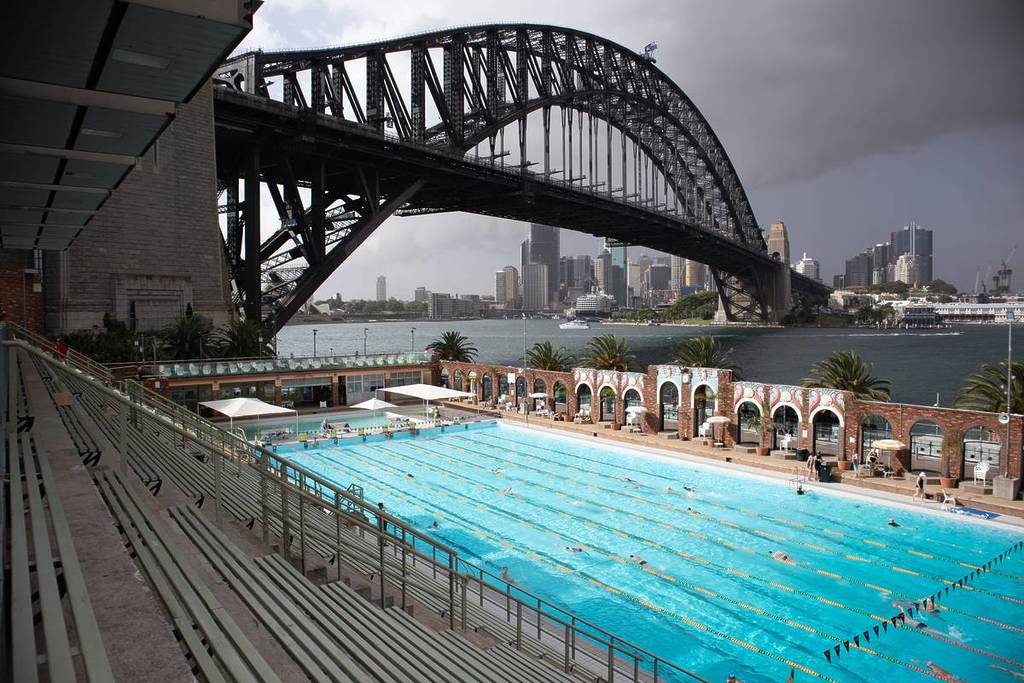 North Sydney Olympic Pool beneath the Sydney Harbour Bridge with swimmers in the 50-metre pool and the city skyline in the background