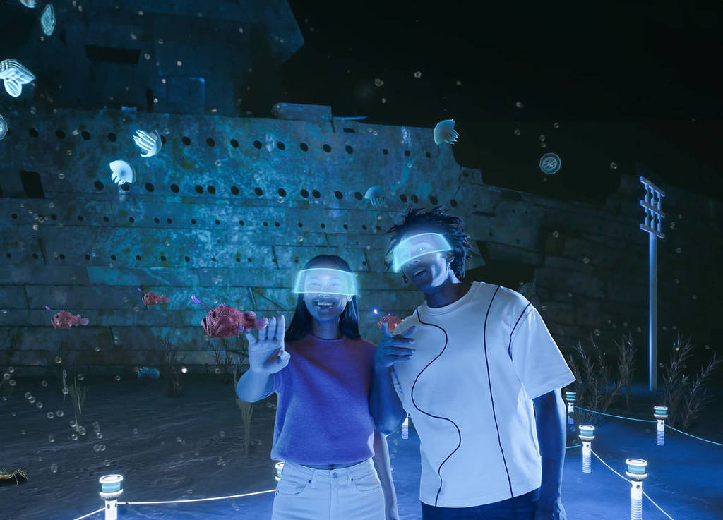 A young man and woman with VR headsets reach out to touch some fish. They appear to be at the bottom of the ocean with the remains of the Titanic behind them.