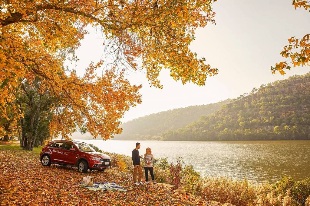 Couple standing by the Hawkesbury River under vibrant autumn foliage at Cattai National Park, with fallen leaves covering the ground and a red car parked nearby