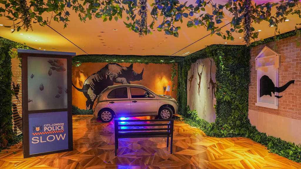 A rhinoceros painted on a wall appears to be climbing on top of a silver car that is parked next to the wall at Banksy: Limitless.