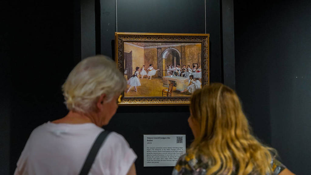 Two women look at the painting, "Simon Cowell Judges the Ballet" at Banksy Limitless.