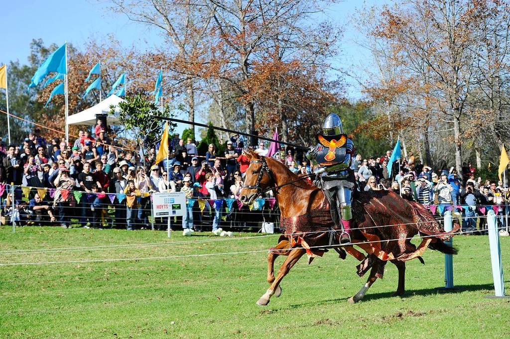 Armoured knight jousting on horseback in front of a large crowd at a medieval festival