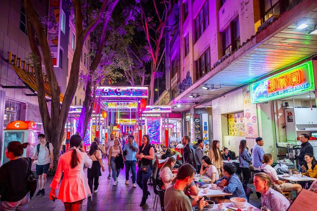 Busy Chinatown street at night with neon signage, outdoor dining and crowds walking through a brightly lit laneway