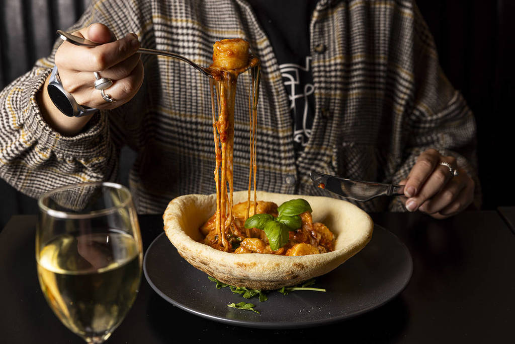 Close-up of cheesy pasta being lifted from a bowl, with melted cheese stretching and a glass of white wine on the table