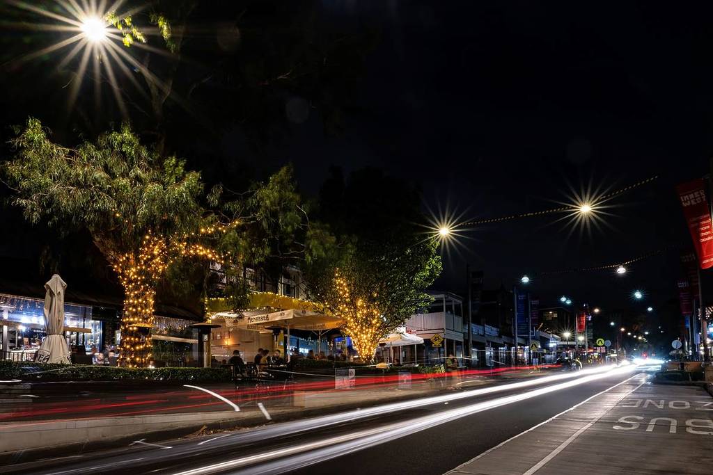 Norton Street in Leichhardt at night with lit-up trees, outdoor dining and traffic passing through the suburb