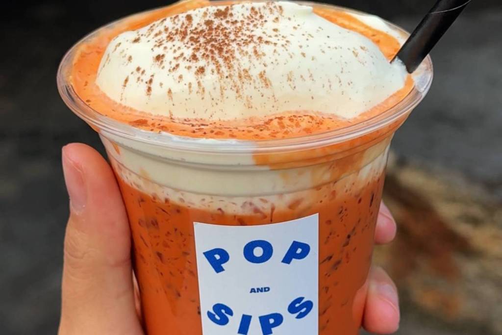 A hand holds a bright orange Southern Thai iced tea from Pop & Sips, topped with a thick layer of soft cream and a dusting of cinnamon, served in a clear takeaway cup