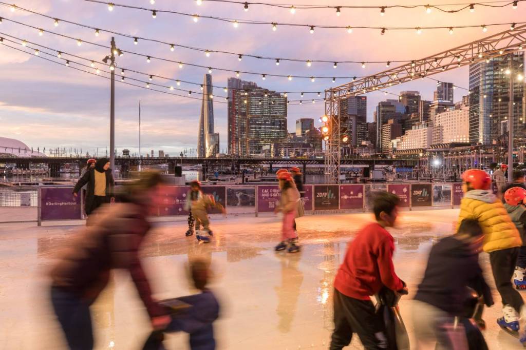 People ice skating at Darling Harbour's outdoor rink at sunset, with city lights and the Sydney skyline in the background