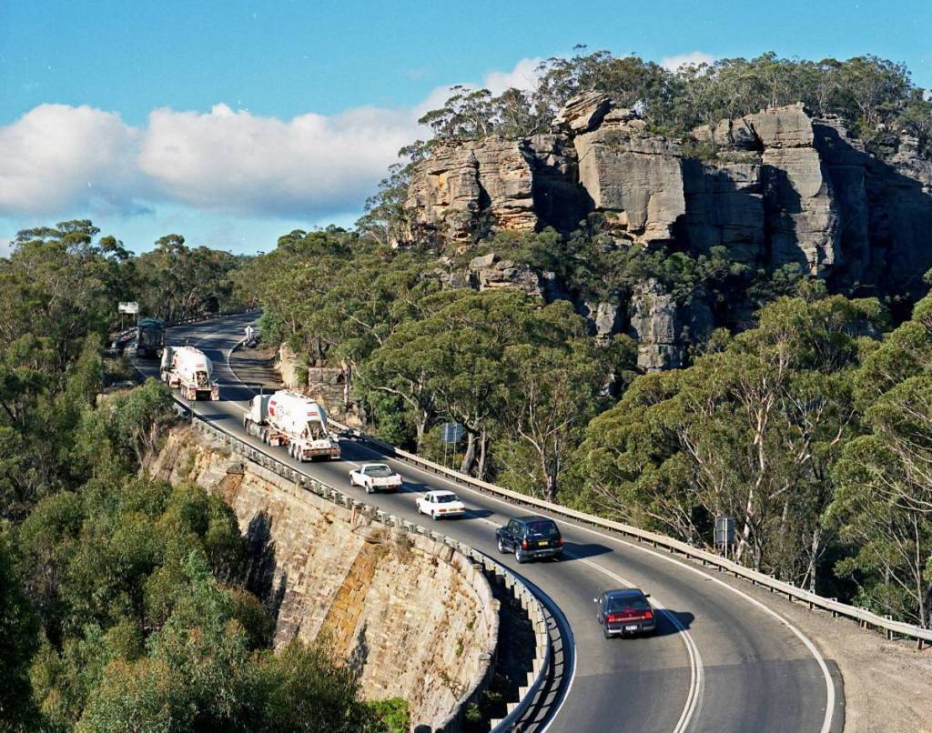 Vehicles travel along the Great Western Highway at Victoria Pass, winding through sandstone cliffs and bushland in the Blue Mountains