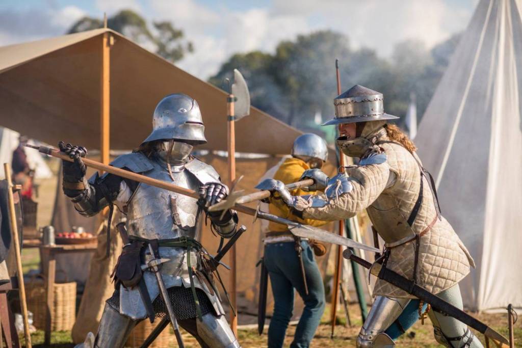 Armoured medieval re-enactors take part in a live combat demonstration at Winterfest Sydney Medieval Fair, with tents and historical encampments in the background.
