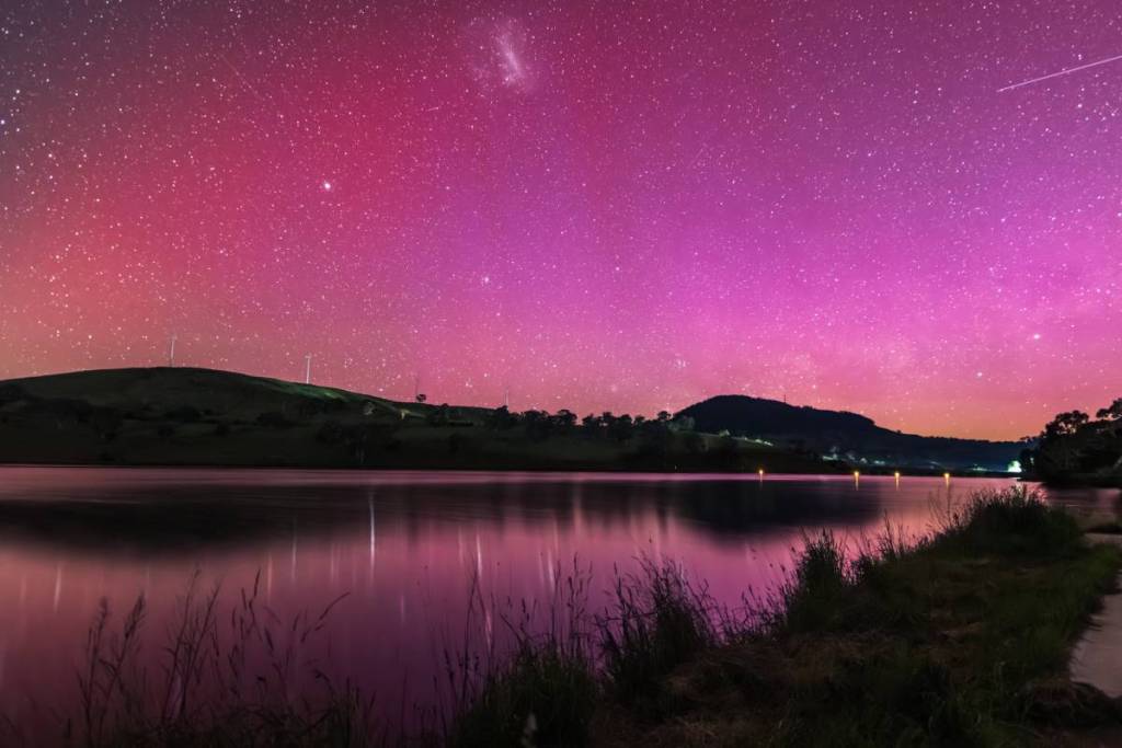 The Aurora Australis illuminates the night sky in vivid shades of pink and purple over a calm lake in regional NSW
