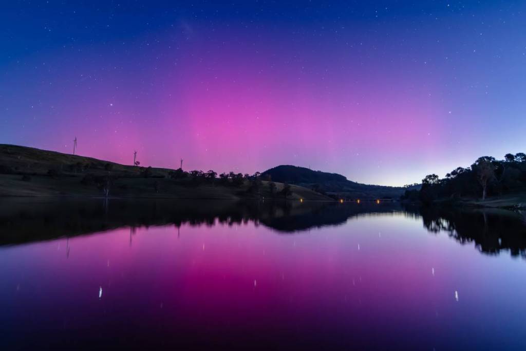 The Aurora Australis creates a pink and purple glow above a tranquil lake as stars shine across the night sky in regional NSW.