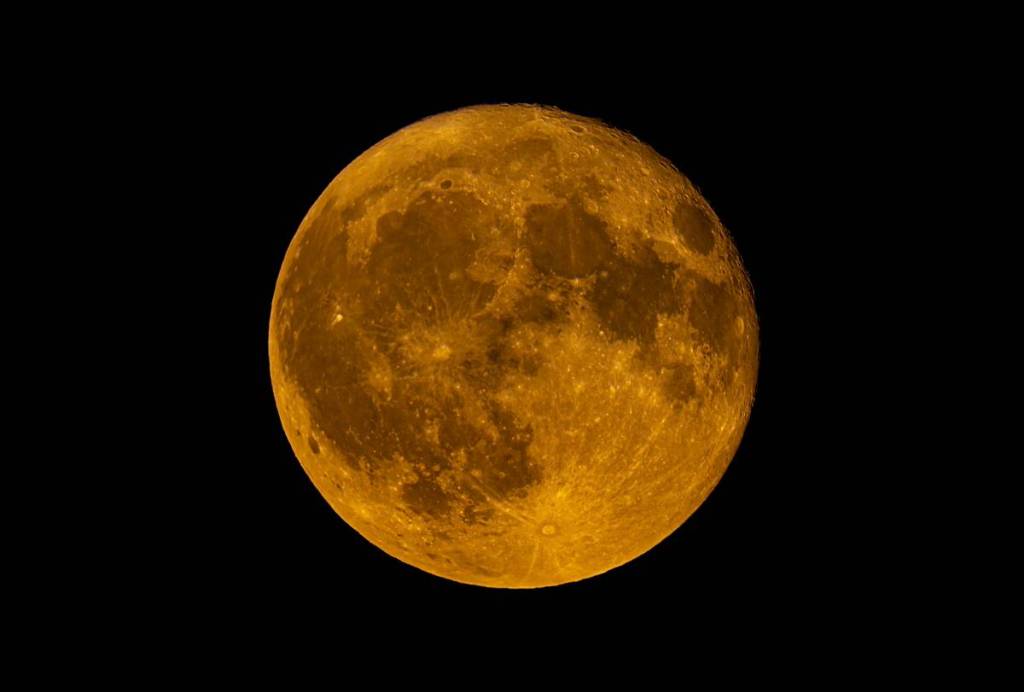 A golden full moon rising against a black night sky, illuminating its craters and lunar surface