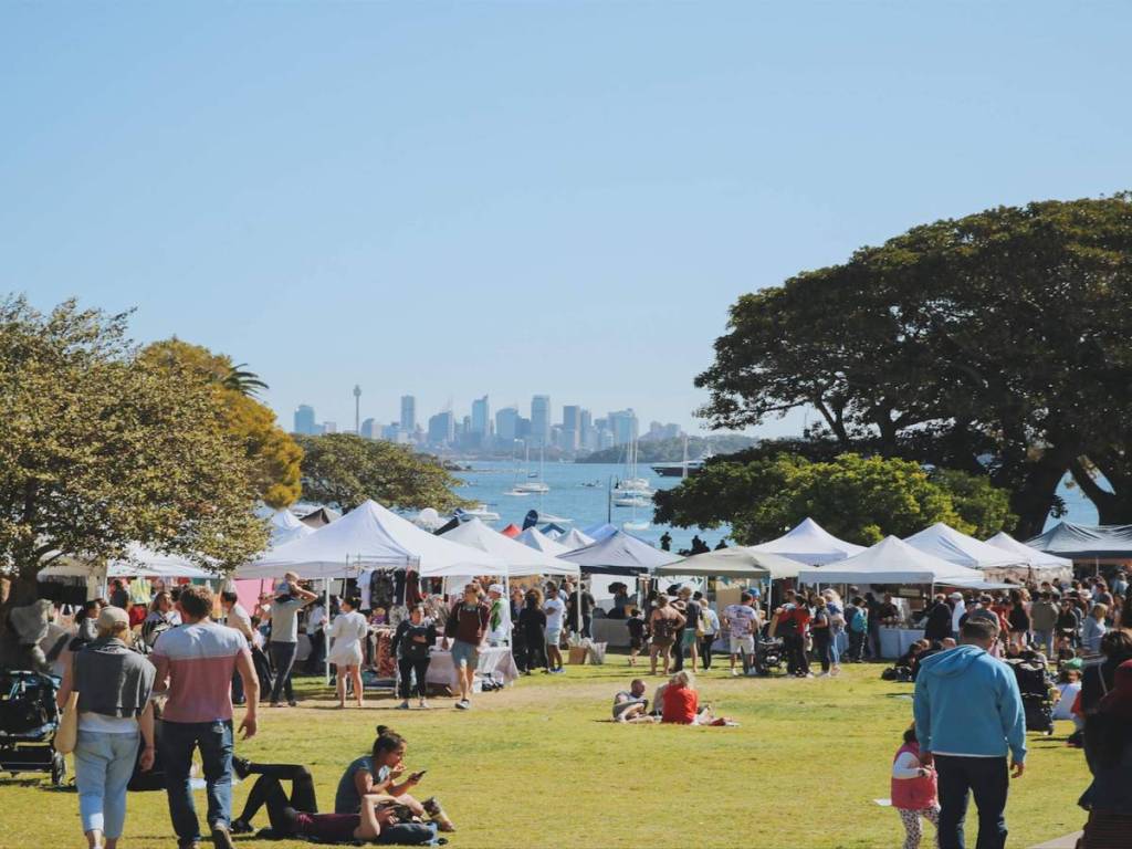 Crowds browsing outdoor market stalls at Watsons Bay, with Sydney Harbour and the city skyline in the background