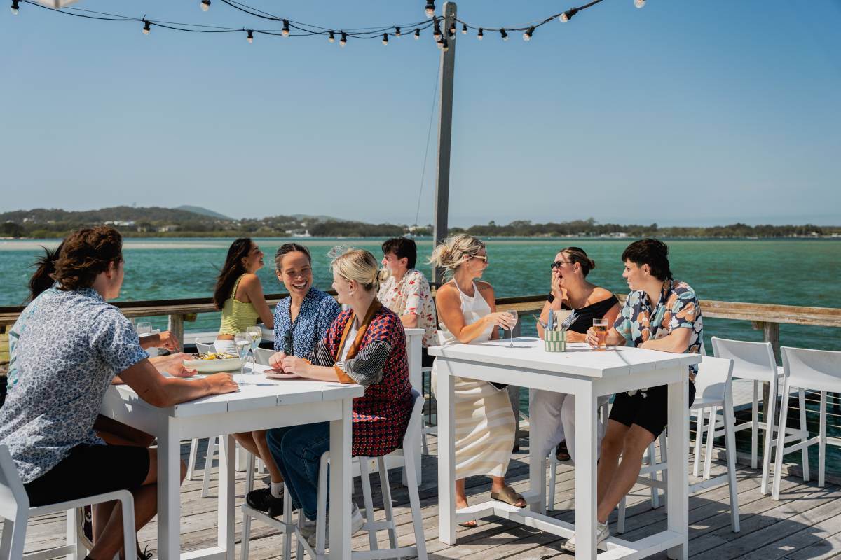 A group of friends dine outdoors at a waterfront restaurant overlooking Wallis Lake