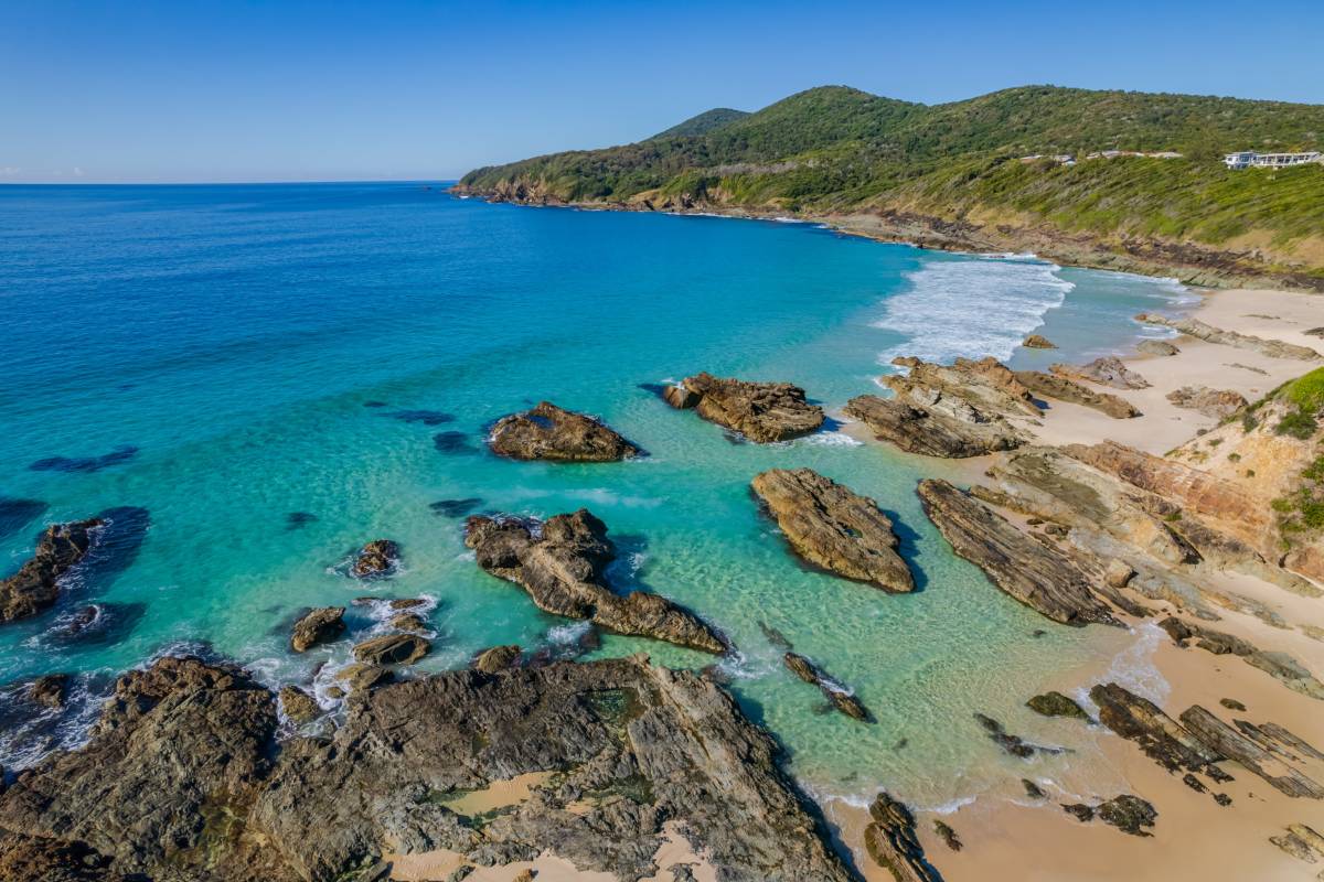 Crystal-clear turquoise water surrounds rocky outcrops and golden sand at Burgess Beach in Forster