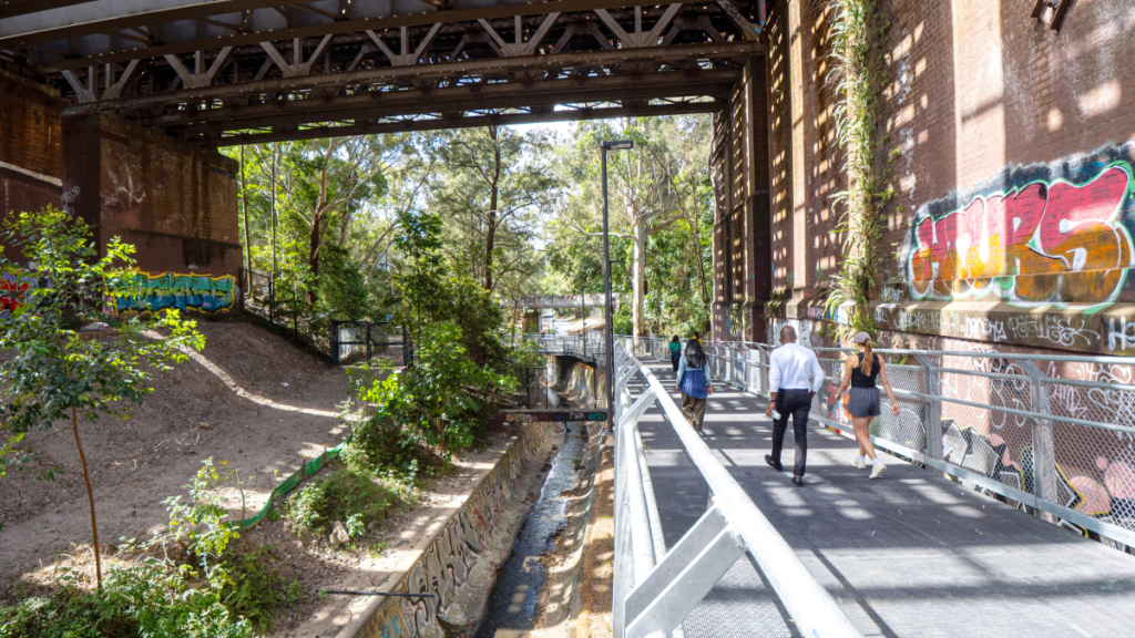 People walking along the GreenWay beneath a railway bridge in Sydney's Inner West, surrounded by trees and colourful street art