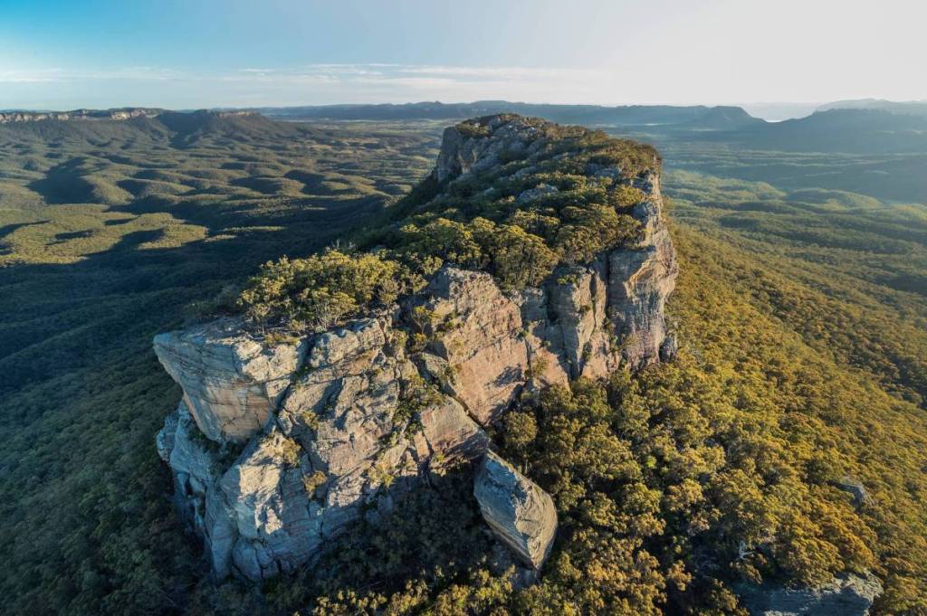 Aerial view of a towering sandstone rock formation surrounded by forest and distant mountain ranges in Capertee Valley
