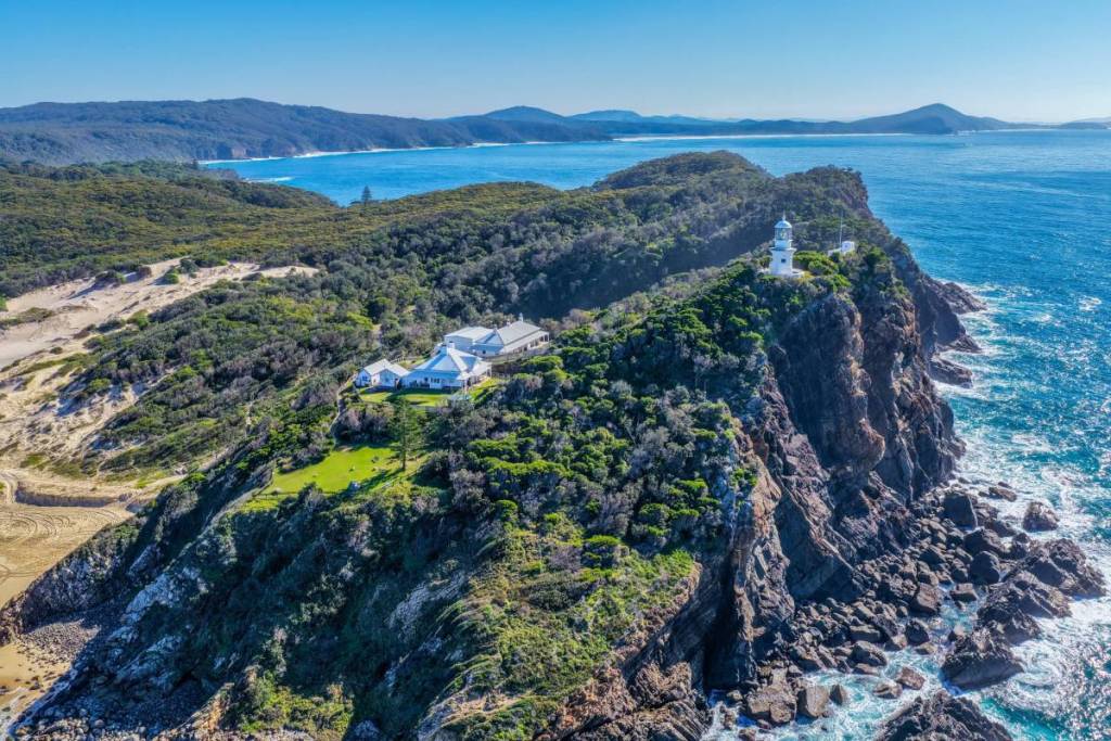 Aerial view of Sugarloaf Point Lighthouse perched on a rugged headland, surrounded by blue ocean and forested coastline at Seal Rocks