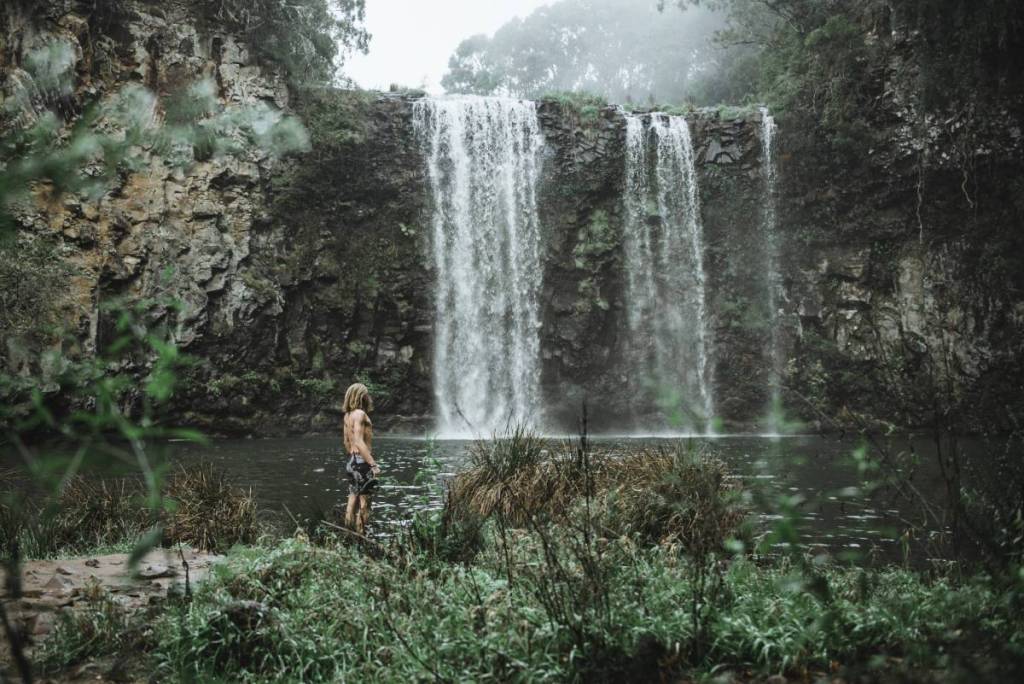 A person standing beside a large waterfall cascading over a rocky cliff into a natural pool, surrounded by lush greenery near Dorrigo, NSW