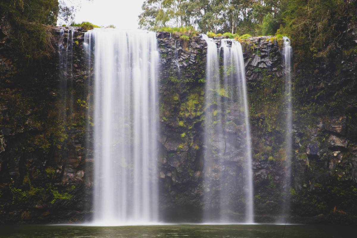 A powerful waterfall cascading over a wide rocky cliff into a natural pool, surrounded by trees and greenery in NSW