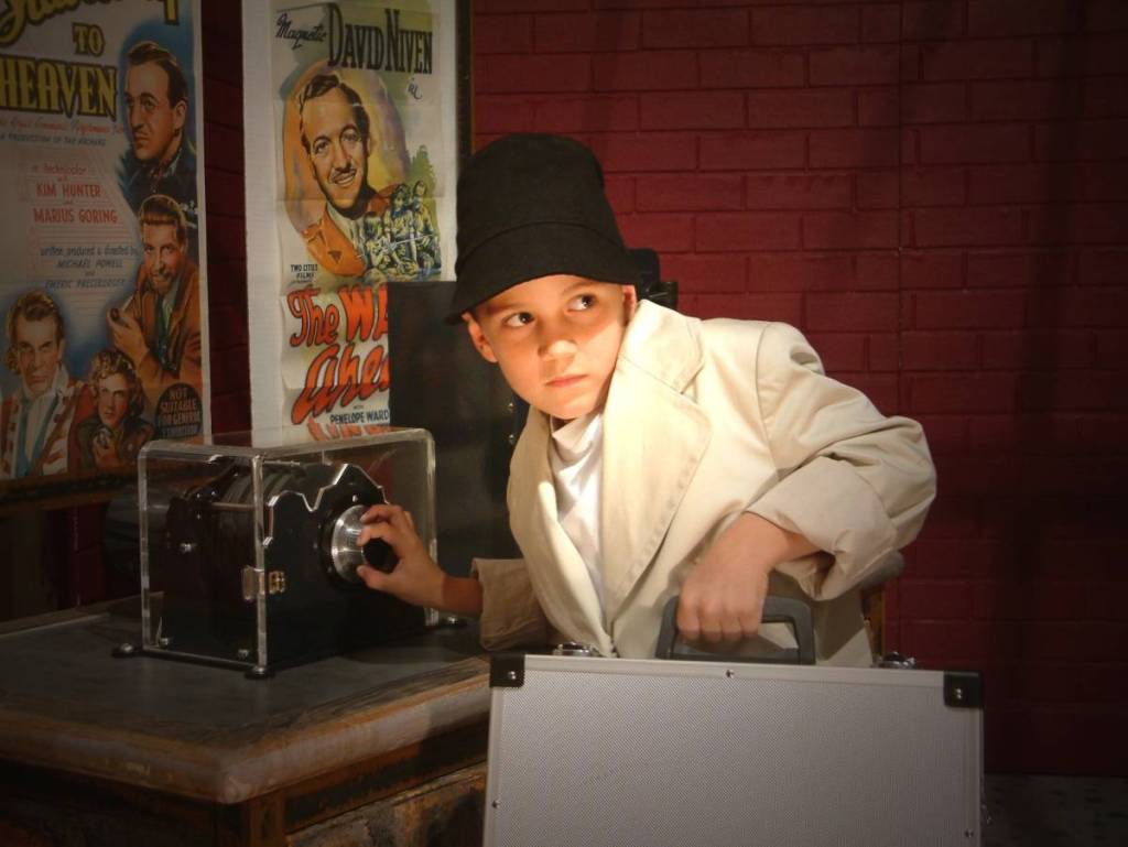 A young boy in a detective's hat and trench coat holds a briefcase at Top Secret: License to Spy. 