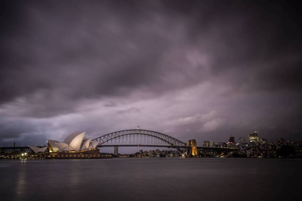 Dark storm clouds loom over Sydney Harbour, with the illuminated Opera House and Harbour Bridge beneath an overcast sky