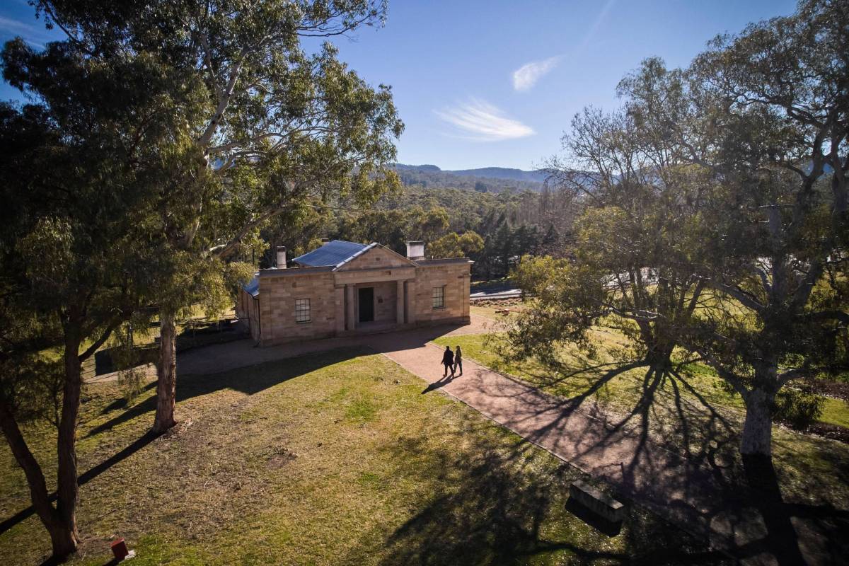 Two visitors walk towards a historic sandstone building surrounded by eucalyptus trees and mountain scenery in Hartley, NSW