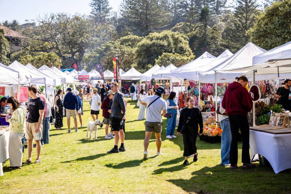 Shoppers browsing rows of white market stalls on a sunny day, with handmade goods, clothing and accessories on display at an outdoor Cambridge Markets event in Sydney