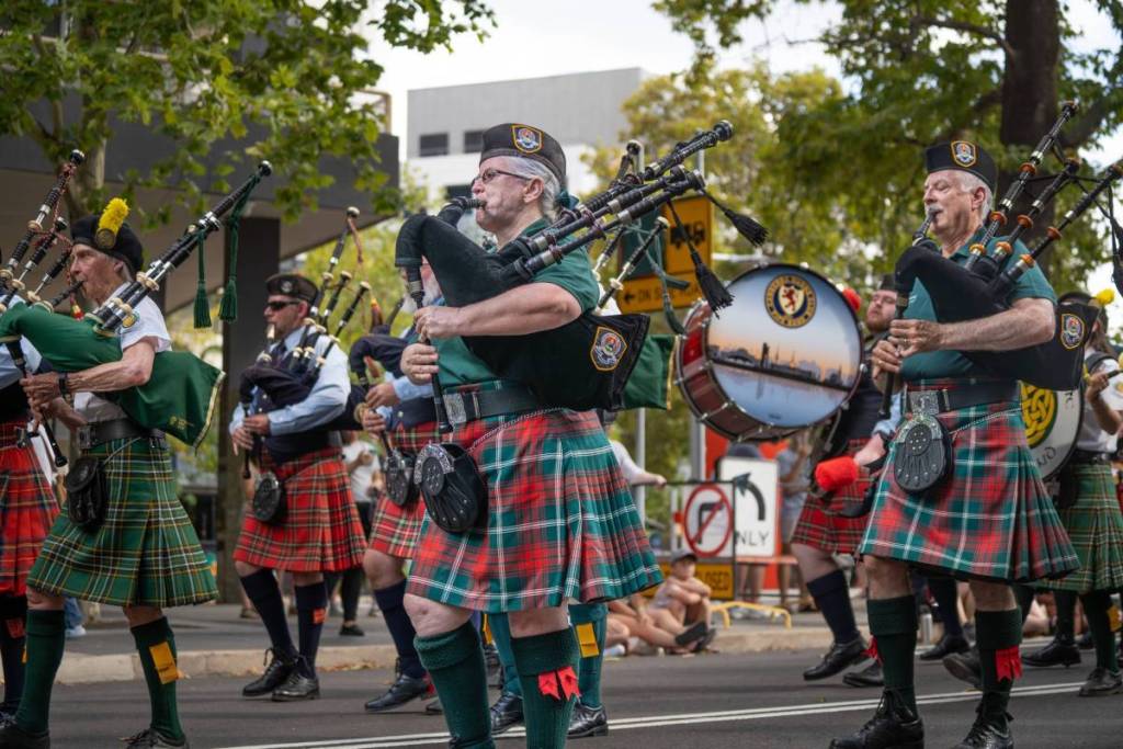 Scottish pipe band members dressed in green and red tartan kilts play bagpipes while marching through Canberra's Multicultural Festival