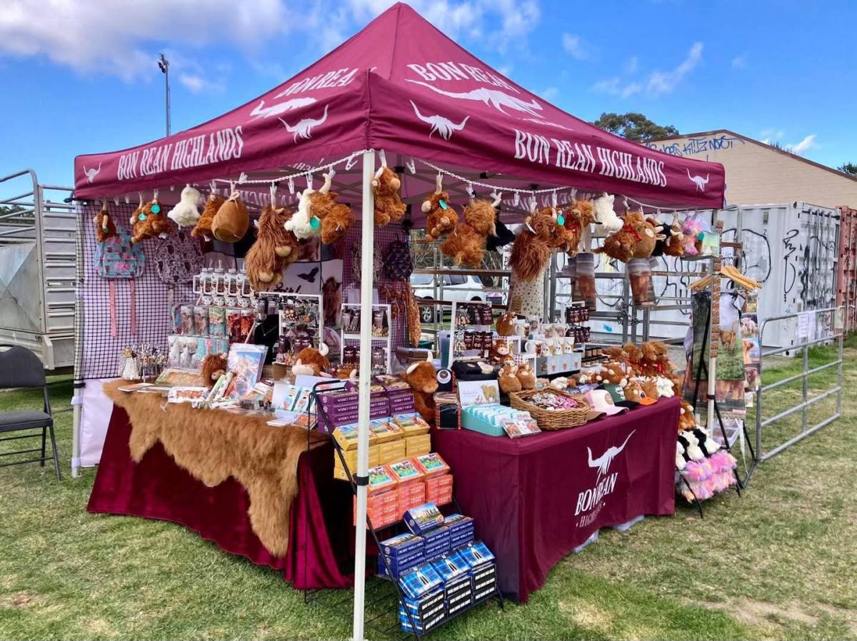 Scottish Highland-themed market stall displaying Highland cow plush toys, souvenirs, clothing and gifts beneath a burgundy marquee