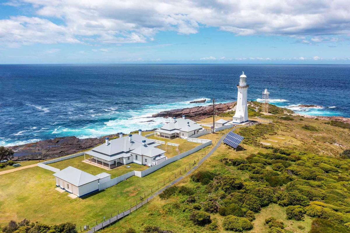 Aerial view of Green Cape Lighthouse and its historic white cottages on a rugged coastal headland, surrounded by blue ocean and crashing waves in Beowa National Park, NSW