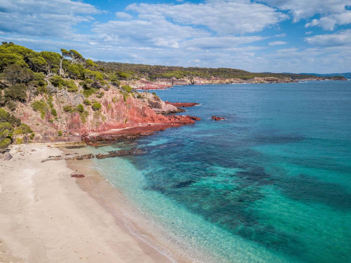 Aerial view of a secluded beach with turquoise waters, red sandstone cliffs and coastal bushland along the NSW South Coast
