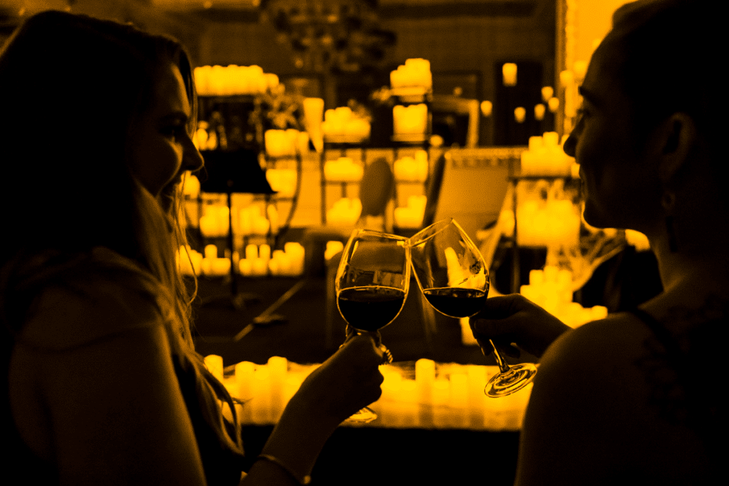 A couple toasting with glasses of wine with the glow of candlelight behind them lighting up a stage set up for musicians.