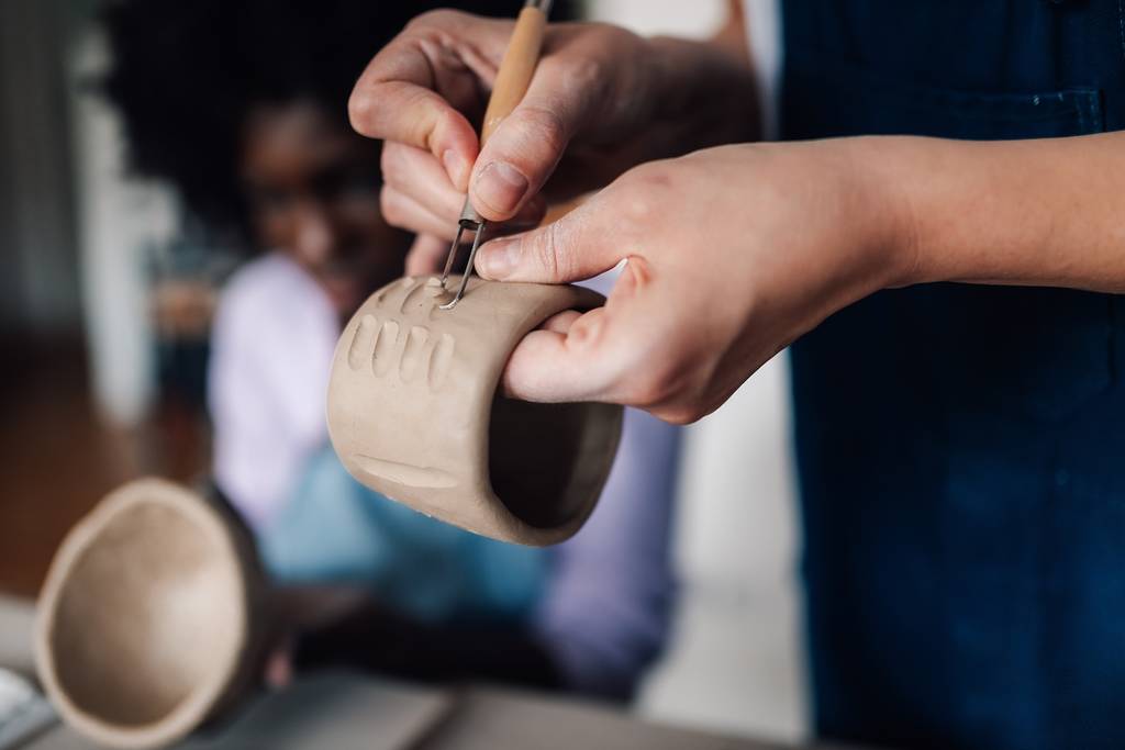 Close up of pottery class tutor standing at studio with clay work in hands and decorating pottery while showing technique to her students. Cropped picture of a pottery course teacher decorating pottery.