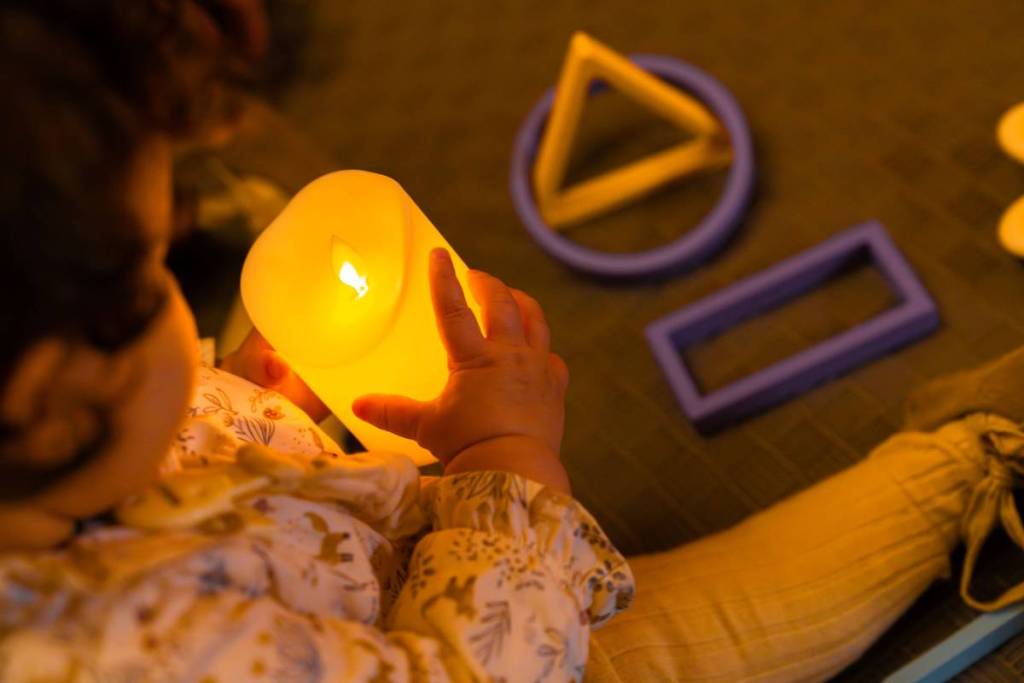 Kind sitzt bei "Candlelight Babies" auf dem Boden und hält eine LED-Kerze in der Hand, daneben liegen Spielzeuge