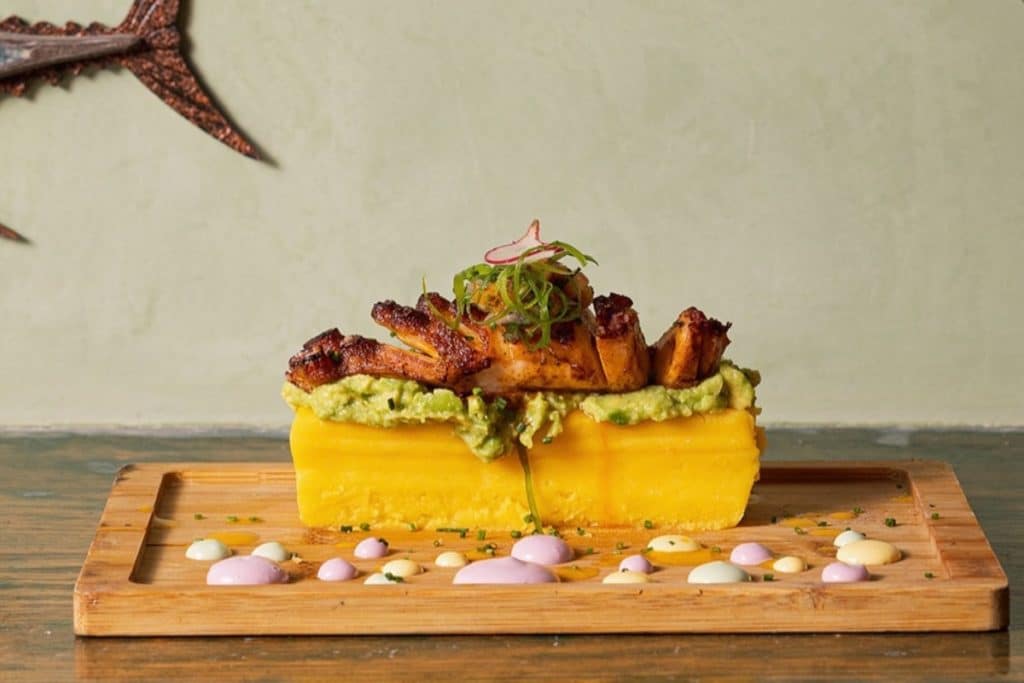 Masa de causa limeña rellena de aguacate con emulsión de ají amarillo y pulpo anticuchero con salsa tártara