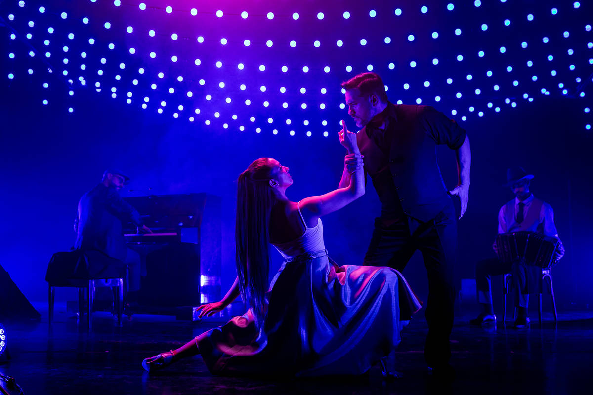 Dos bailarines en un escenario iluminado de guirnaldas azules bailando tango y acompañados por un pianista y bandoneonista