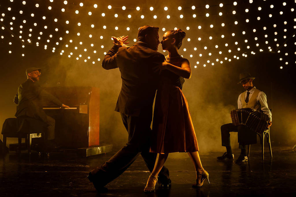 Dos bailarines de tango en un escenario lleno de guirnaldas y con un pianista y un bandoneonista