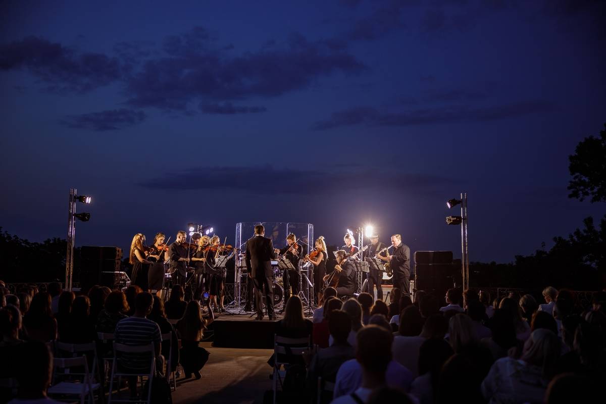 Orquesta de cámara interpretando música clásica al aire libre al caer la noche, con el público sentado y luces sobre el escenario