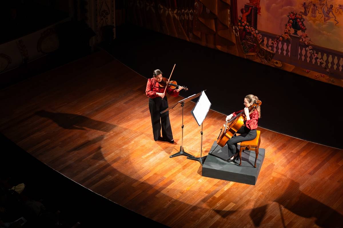 Dúo de violinista y chelista actuando en un teatro con suelo de madera y decorado clásico, bajo la luz de los focos.