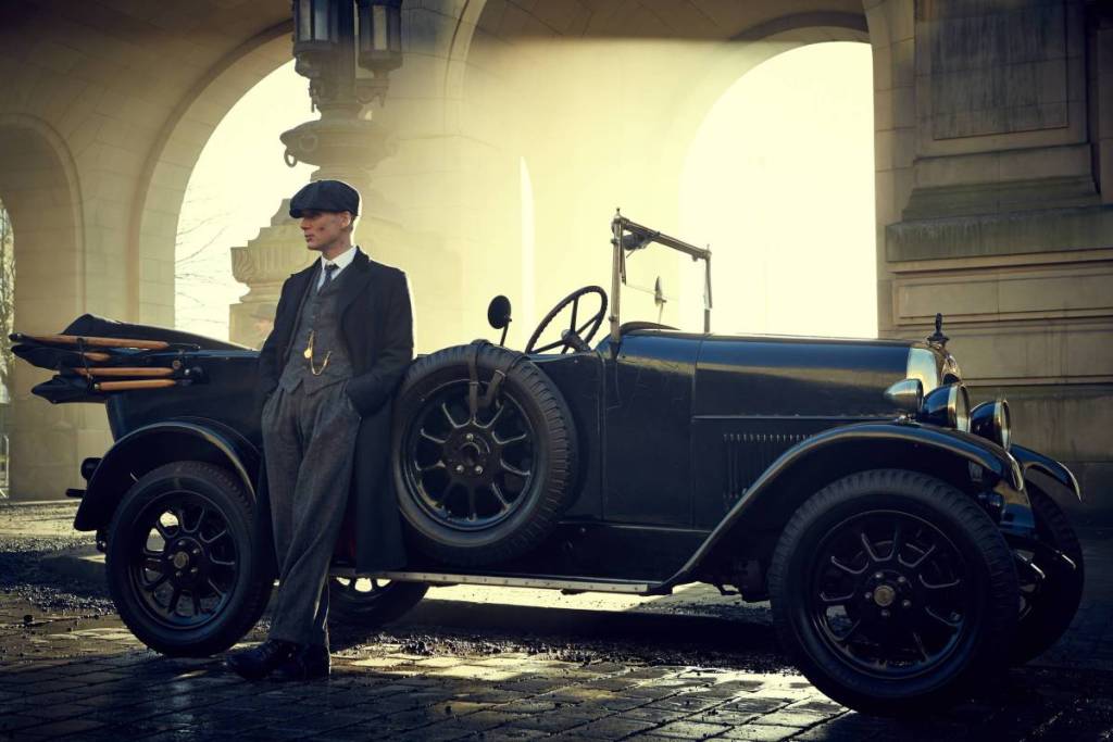 A wide shot of Tommy Shelby waiting in front of a vintage car in a scene from *Peaky Blinders*.