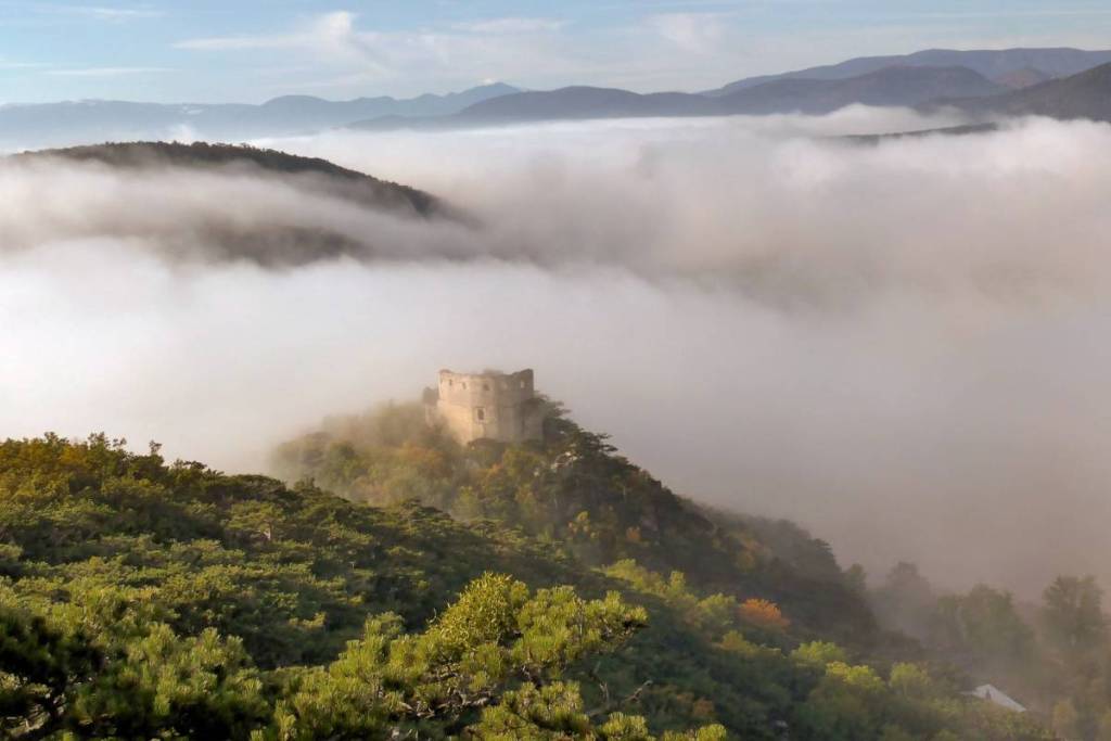 Die Burgruine Emmerberg thront auf einem bewaldeten Hügel, der majestätisch aus einem dichten Nebelmeer im Tal herausragt, mit weiteren Bergzügen im Hintergrund.
