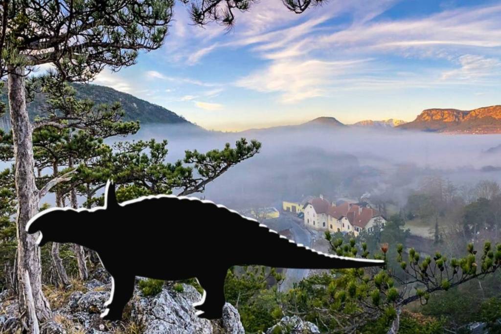Schwarze Silhouette des gepanzerten Zwergdinosauriers Struthiosaurus austriacus auf einem Felsen im Vordergrund, mit Blick auf das nebelverhangene Muthmannsdorf und die sonnenbeschienene Hohe Wand im Hintergrund.