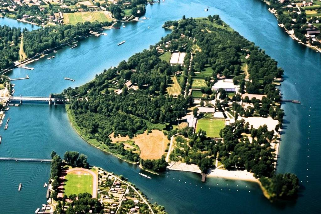 Luftaufnahme der dicht bewaldeten Insel Gänsehäufel, die wie eine grüne Oase inmitten des blauen Wassers der Alten Donau in Wien liegt.