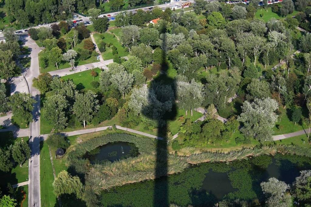 Luftbild des grünen Donauparks mit Spazierwegen und einem kleinen Teich, über den sich der markante, riesige Schatten des Donauturms legt.