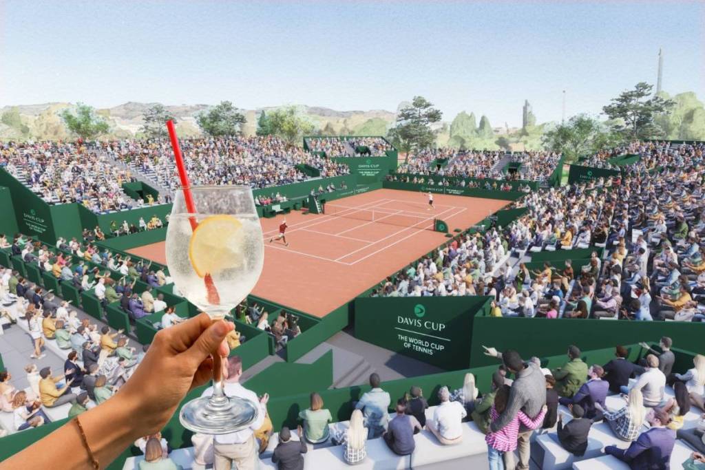 A close-up of a hand holding a glass of spritzer with a straw. In the background is a packed Davis Cup pop-up stadium in Vienna featuring a clay court, lots of spectators, and a large “DAVIS CUP” sign outdoors.