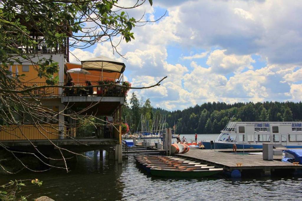 Das Seerestaurant Ottenstein mit seiner gut besuchten Sonnenterrasse direkt am Wasser. Davor liegt der Bootsverleih mit Holzruderbooten und einem Ausflugsschiff, die an Holzstegen vertäut sind.